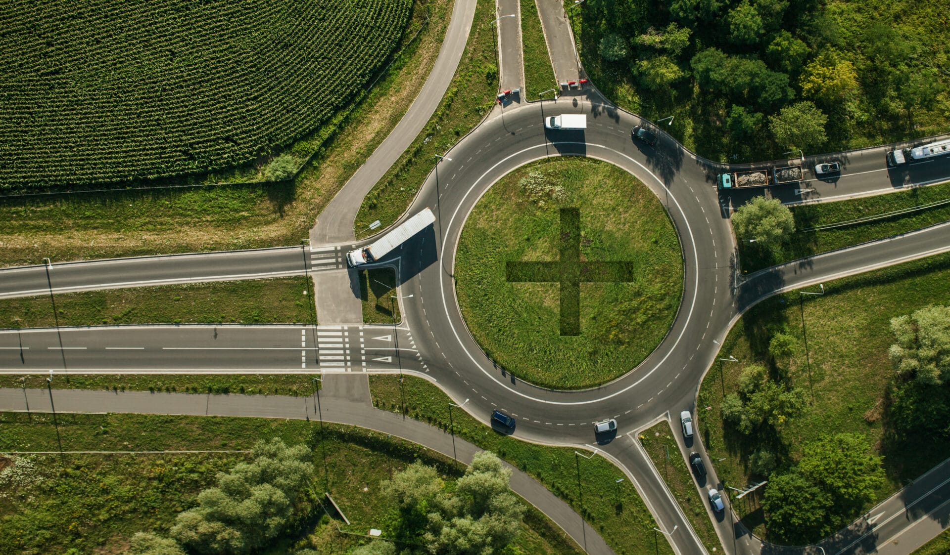 Aerial view of a roundabout with a grassy center shaped like a plus sign. multiple vehicles, including a truck and cars, navigate the roundabout surrounded by greenery.