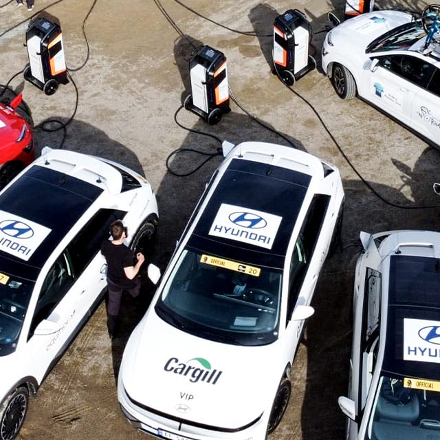 Overhead view of several white Hyundai electric cars parked in a charging area, each connected to a charging station. One person is seen near a car, and the ground is wet, suggesting recent rain. The cars have "Hyundai" logos and other branding, including "Cargill VIP".