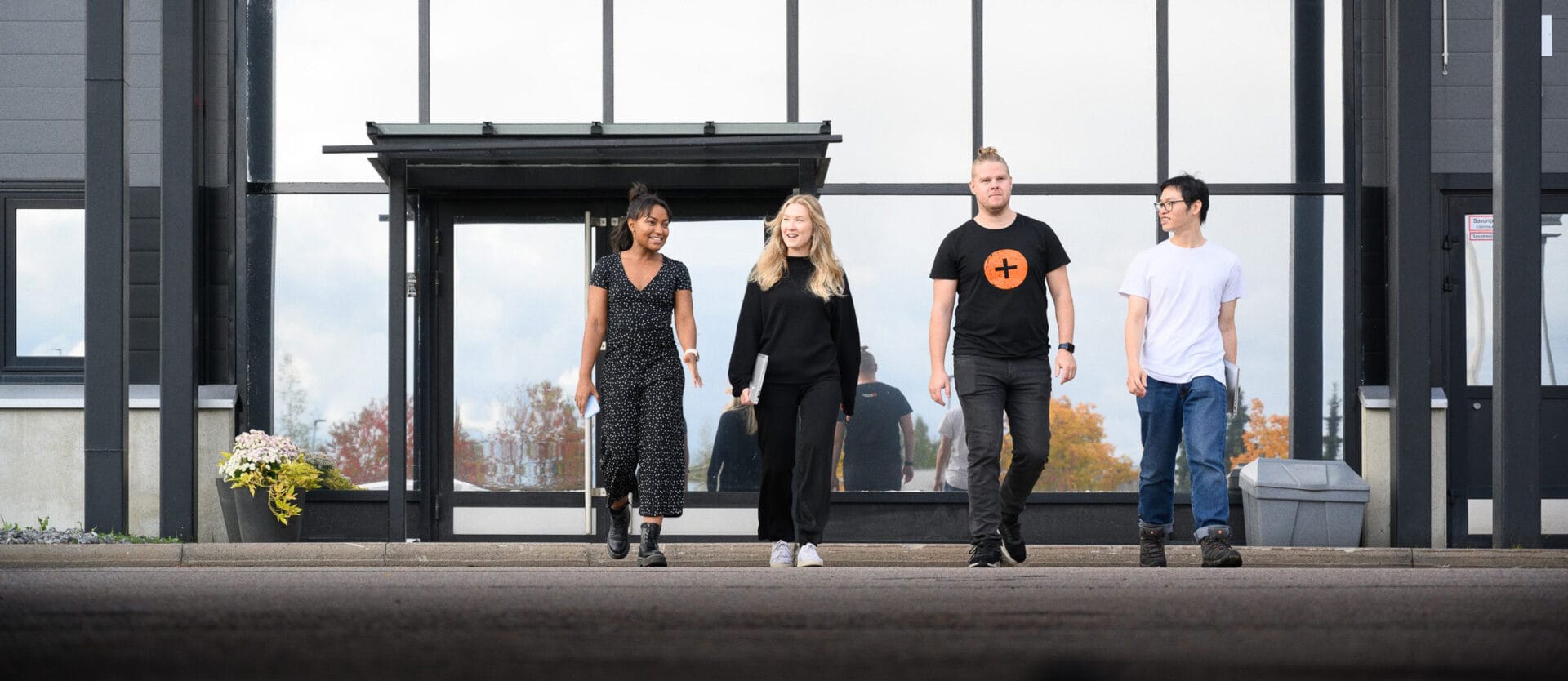 Four people walking side by side outside a modern glass building. From left to right: a woman in a black dress, a woman in black pants and top, a man in a black t-shirt with orange cross and jeans, and a man in a white t-shirt and jeans. The background shows autumn trees.