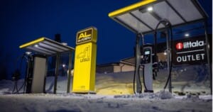 Electric vehicle charging station lit up at night, surrounded by snow. A yellow AL Station sign is in the center, with charging cables and a nearby building displaying "iittala OUTLET.
